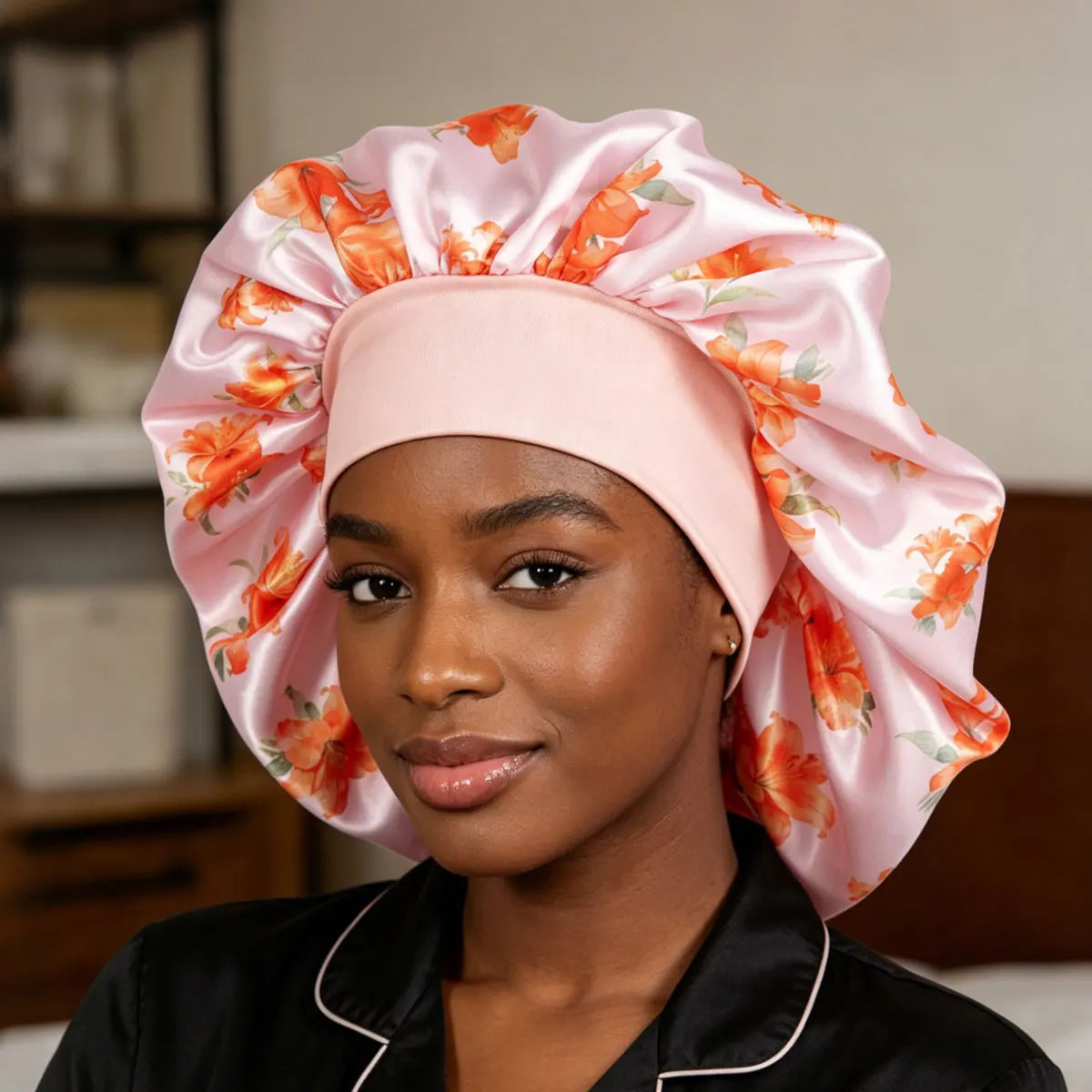 A woman wearing a light pink satin sleep cap with an orange floral print, paired with a black satin pajama top with white piping.