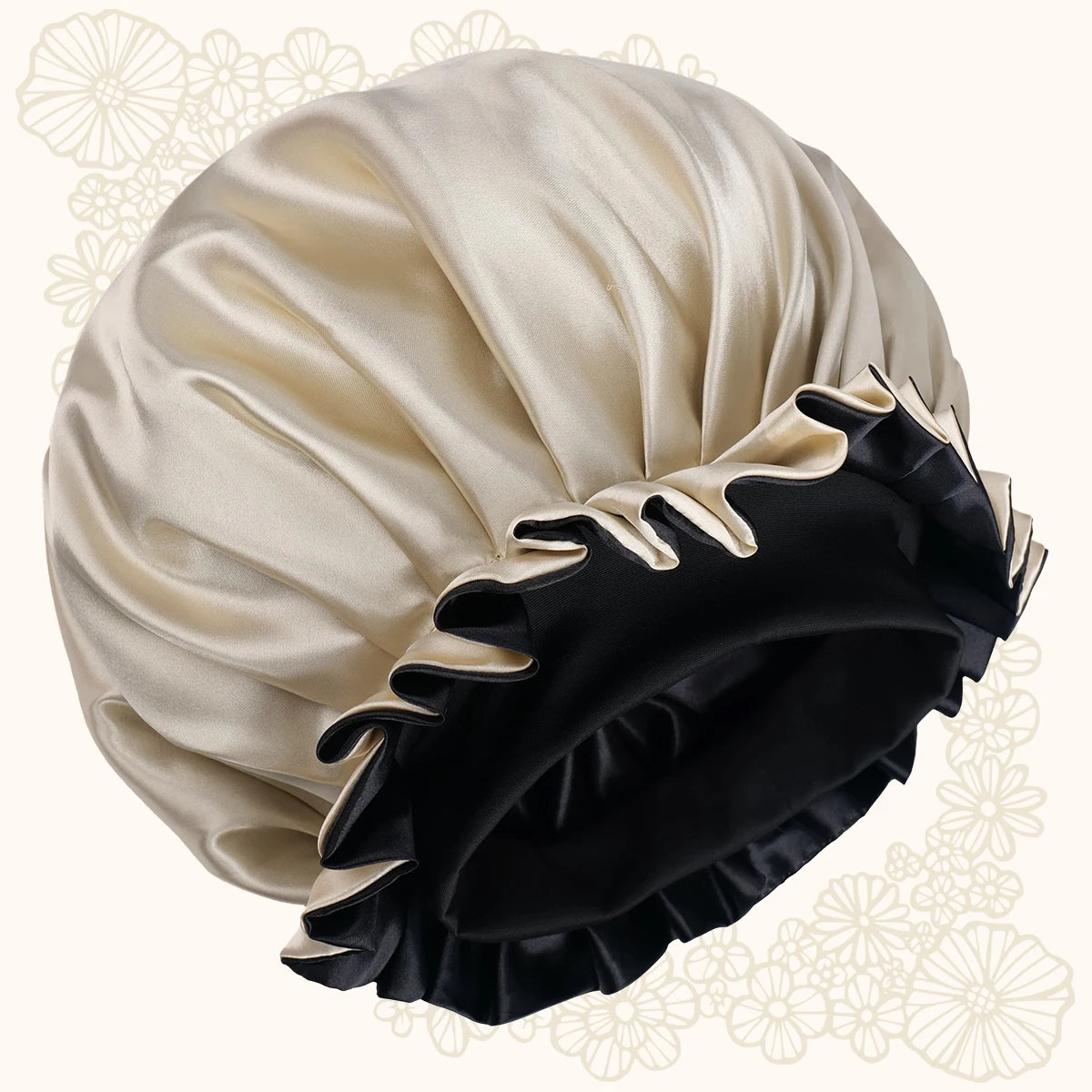 A satin shower cap in cream and black, featuring a pleated cream exterior and a black ruffled trim with cream piping, set against a light cream background with faint floral patterns.